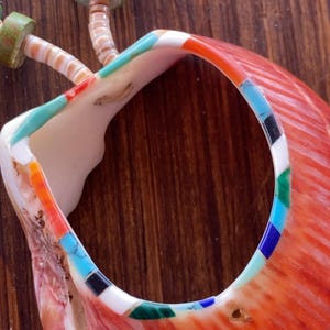 May include: Close-up of a handcrafted necklace featuring a large, vibrant orange shell pendant with a circular opening. The pendant is adorned with inlaid turquoise, black, white, and green stones. The necklace includes green and turquoise beads.
