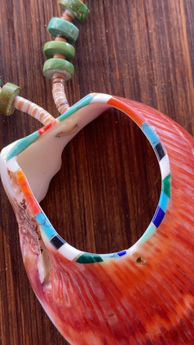 May include: Close-up of a handcrafted necklace featuring a large, vibrant orange shell pendant with a circular opening. The pendant is adorned with inlaid turquoise, black, white, and green stones. The necklace includes green and turquoise beads.
