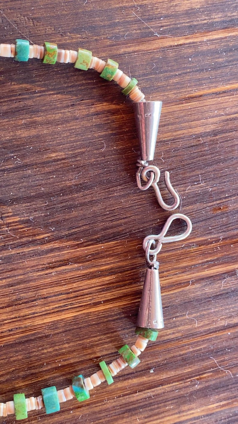 May include: A close-up of a necklace with a silver clasp. The necklace features small, square, turquoise and green beads interspersed with off-white shell beads. The clasp is a simple hook and eye design.