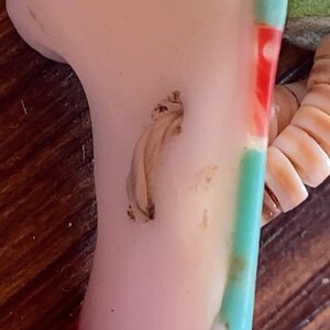 May include: A close-up of a section of a ceramic figurine. The figurine has a pale pink surface with a light brown, textured detail. A section of the figurine is painted with red, white, and turquoise stripes. The figurine is on a wooden surface.