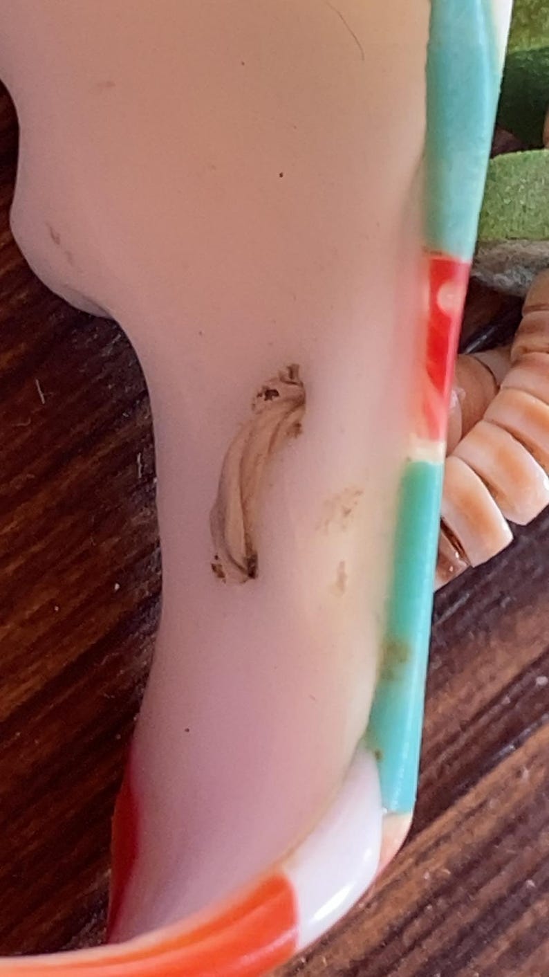 May include: A close-up of a section of a ceramic figurine. The figurine has a pale pink surface with a light brown, textured detail. A section of the figurine is painted with red, white, and turquoise stripes. The figurine is on a wooden surface.