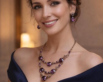 May include: A multi-strand necklace featuring round, deep purple faceted stones set in a metallic bronze-toned chain, paired with matching drop earrings that feature a single round purple stone, worn with a dark navy blue off-the-shoulder dress.