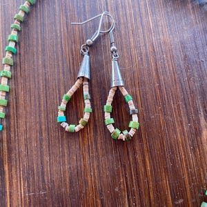 May include: A necklace and matching earrings set. The jewelry features a combination of green, turquoise, and off-white beads. The earrings have silver-colored cone-shaped tops. The necklace and earrings are displayed on a wooden surface.