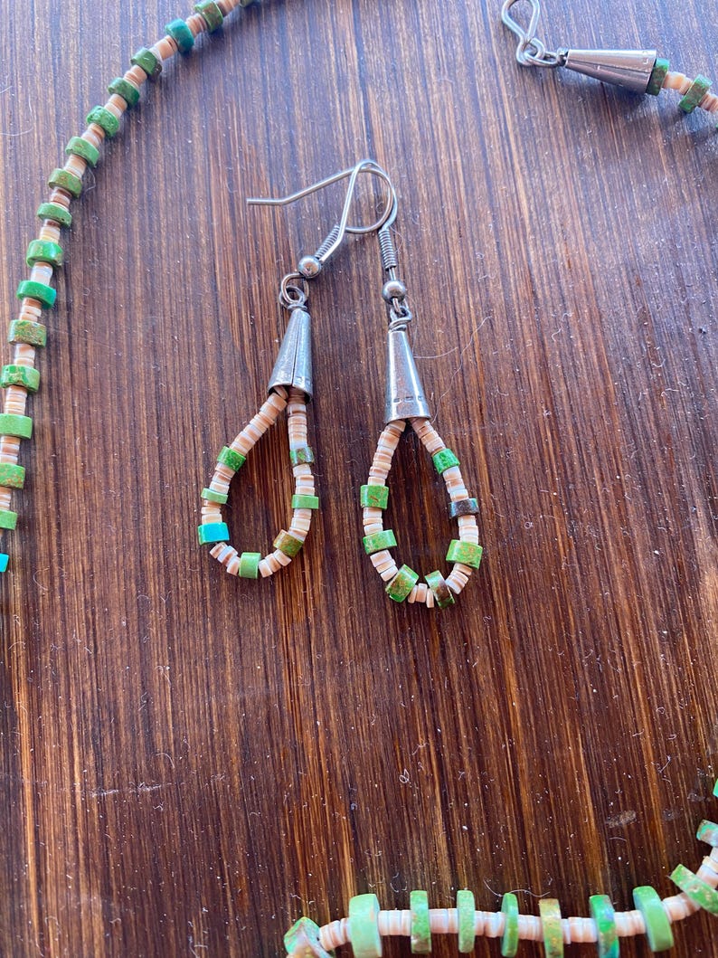 May include: A necklace and matching earrings set. The jewelry features a combination of green, turquoise, and off-white beads. The earrings have silver-colored cone-shaped tops. The necklace and earrings are displayed on a wooden surface.