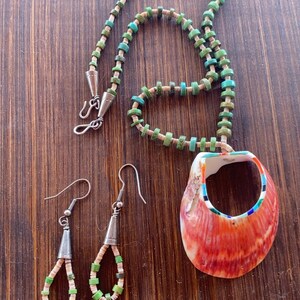 May include: A handcrafted jewelry set featuring a necklace and matching earrings. The necklace has a pendant made from a red and white shell. The necklace and earrings are adorned with turquoise and green beads. The set is displayed on a wooden surface.