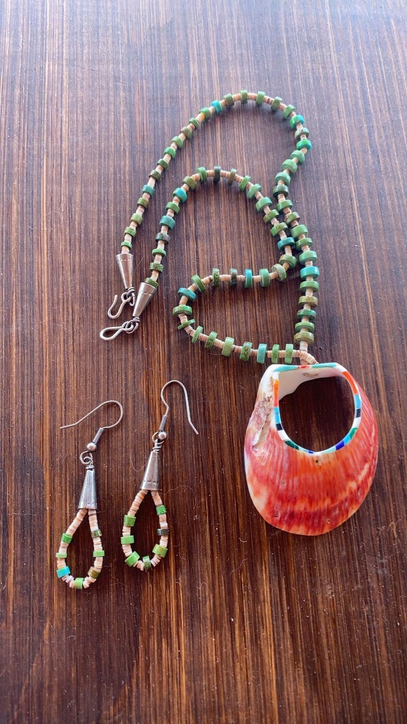 May include: A handcrafted jewelry set featuring a necklace and matching earrings. The necklace has a pendant made from a red and white shell. The necklace and earrings are adorned with turquoise and green beads. The set is displayed on a wooden surface.