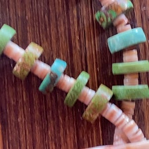 May include: Close-up of a necklace with a seashell pendant. The necklace features light green and turquoise-colored beads strung on a beige cord. The seashell pendant has a white and orange interior. The background is a dark brown wooden surface.