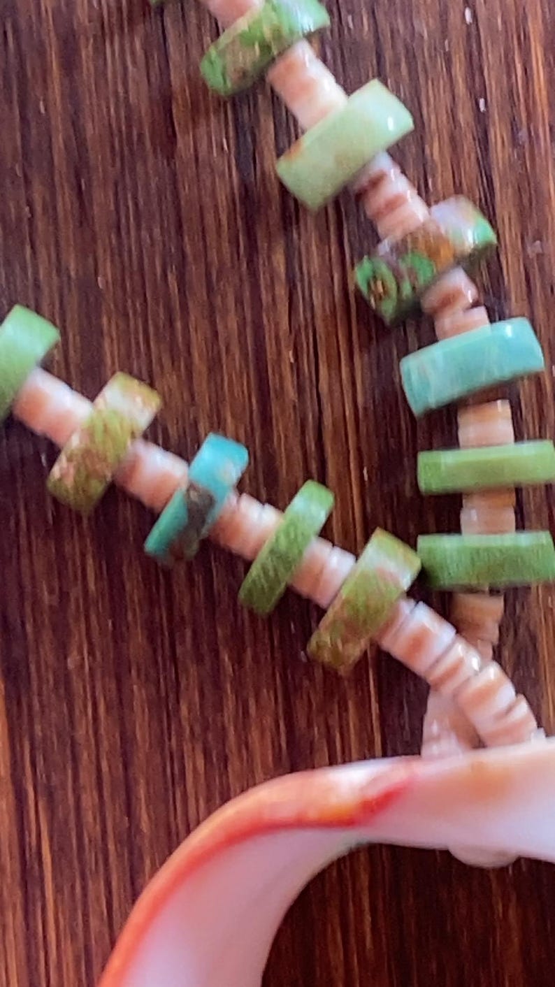 May include: Close-up of a necklace with a seashell pendant. The necklace features light green and turquoise-colored beads strung on a beige cord. The seashell pendant has a white and orange interior. The background is a dark brown wooden surface.