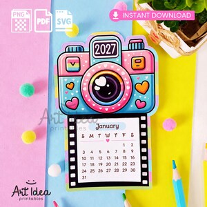 Può includere: Un calendario 2027 con un'illustrazione colorata di una fotocamera nei toni del rosa e del blu, dettagli a forma di cuore e un bordo a pellicola. La pagina mostra il mese di gennaio con una griglia di date in uno stile grafico giocoso.