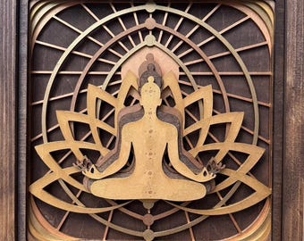 May include: A wooden wall art piece featuring a layered, intricate design of a person meditating in a lotus position. The artwork is framed in a dark wood frame and has a geometric pattern in the background.