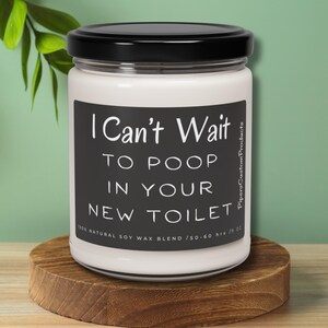 May include: A white candle in a glass jar with a black lid. The label reads "I Can't Wait to Poop in Your New Toilet." The candle sits on a small, round wooden stand.