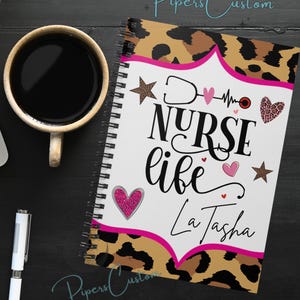 May include: A spiral notebook with a leopard print cover. The front of the notebook has a white background with the text "Nurse life" in black and pink lettering. The text "La Tasha" is also in black and pink lettering. The notebook has pink hearts, stars, and a stethoscope graphic.