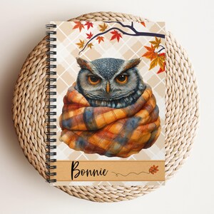 May include: Spiral-bound notebook featuring a detailed illustration of an owl wrapped in a plaid scarf. The background has a beige plaid pattern with autumn leaves and a branch. The name "Bonnie" is written on a wooden banner.
