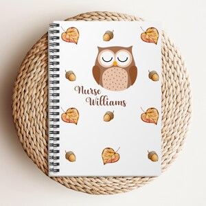 May include: A spiral-bound notebook with a white cover featuring a brown owl illustration, acorns, and autumn leaves. The text "Nurse Williams" is written in brown script. The notebook is resting on a woven, circular placemat.