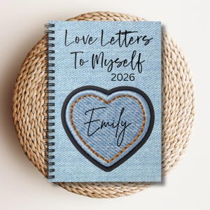 May include: A spiral-bound notebook with a denim-effect cover. The cover features the text "Love Letters To Myself 2026" and a heart-shaped patch with the name "Emily" in cursive. The notebook is resting on a woven, circular placemat.