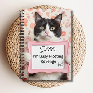 May include: A spiral-bound notebook featuring a black and white cat holding a pink framed sign. The sign reads "Shhh... I'm Busy Plotting Revenge Katy" against a floral background. The notebook is set on a woven, circular placemat.