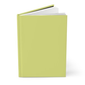May include: A hardcover book with a light green cover and white pages. The book is slightly open, revealing the interior pages. The spine is visible, and the book is set against a white background.