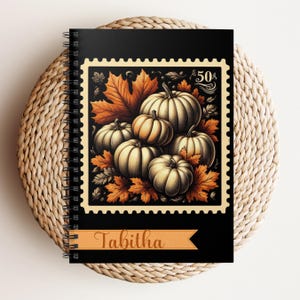 Könnte beinhalten: Ein spiralgebundenes Notizbuch mit schwarzem Einband und einem Vintage-Stempeldesign. Der Stempel zeigt Kürbisse und Herbstblätter in Orange-, Braun- und Cremetönen. Der Name "Tabitha" ist unten auf einem Banner aufgedruckt.