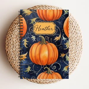 May include: A spiral-bound notebook featuring a fall-themed design. The cover displays orange pumpkins on a dark blue background with gold accents. The name "Heather" is written in a decorative font within a gold frame.