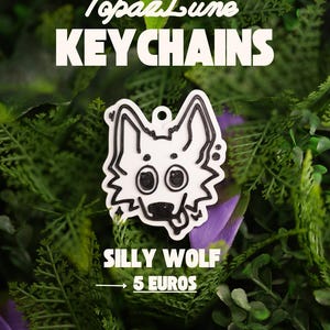 Peut inclure: Un porte-clés blanc avec une illustration au trait noir représentant une tête de loup aux grands yeux avec la langue tirée. Le texte TopazLune KEYCHAINS et SILLY WOLF est imprimé sur l'image. Le motif comporte un trou en haut pour l'attache.