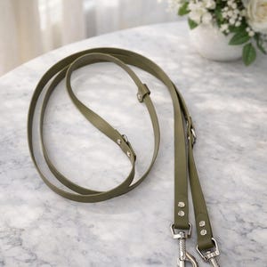 May include: Olive green leather dog lead with silver-coloured metal clasps and hardware. The lead is looped and lies on a marble surface. The lead is designed for pet owners.