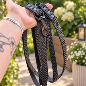 May include: A black dog lead featuring a textured, honeycomb-like surface pattern. The lead includes a matte black metal swivel snap hook and a series of silver-toned rivets along the handle section, providing a secure and durable construction.