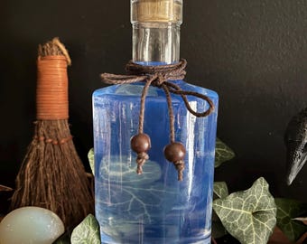 Puede incluir: Una botella de vidrio transparente llena de un líquido azul translúcido. La botella está sellada con un tapón de corcho con un diseño de vid grabado. Un cordel marrón oscuro está atado alrededor del cuello, terminando en dos cuentas de madera.