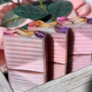 May include: Three bars of handmade soap with a pink base and swirls of gold and purple. The soap is displayed in a wooden frame.