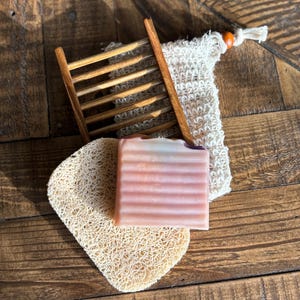 May include: A pink and white bar of soap on a natural loofah sponge, next to a wooden soap dish and a natural fiber washcloth.