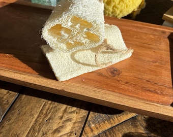 May include: A natural loofah sponge with a white cotton washcloth. The loofah is tied with twine and is on a wooden tray.