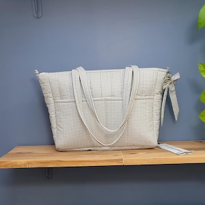 Pode incluir: Uma grande bolsa tote acolchoada com um padr&atilde;o xadrez branco e azul claro. A bolsa tem duas al&ccedil;as longas e v&aacute;rios bolsos externos. Um pequeno acess&oacute;rio correspondente est&aacute; ao lado da bolsa. A bolsa est&aacute; em uma prateleira de madeira.