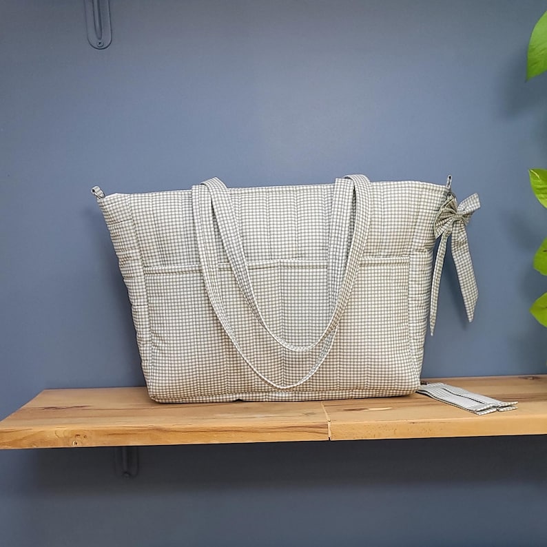 Pode incluir: Uma grande bolsa tote acolchoada com um padr&atilde;o xadrez branco e azul claro. A bolsa tem duas al&ccedil;as longas e v&aacute;rios bolsos externos. Um pequeno acess&oacute;rio correspondente est&aacute; ao lado da bolsa. A bolsa est&aacute; em uma prateleira de madeira.
