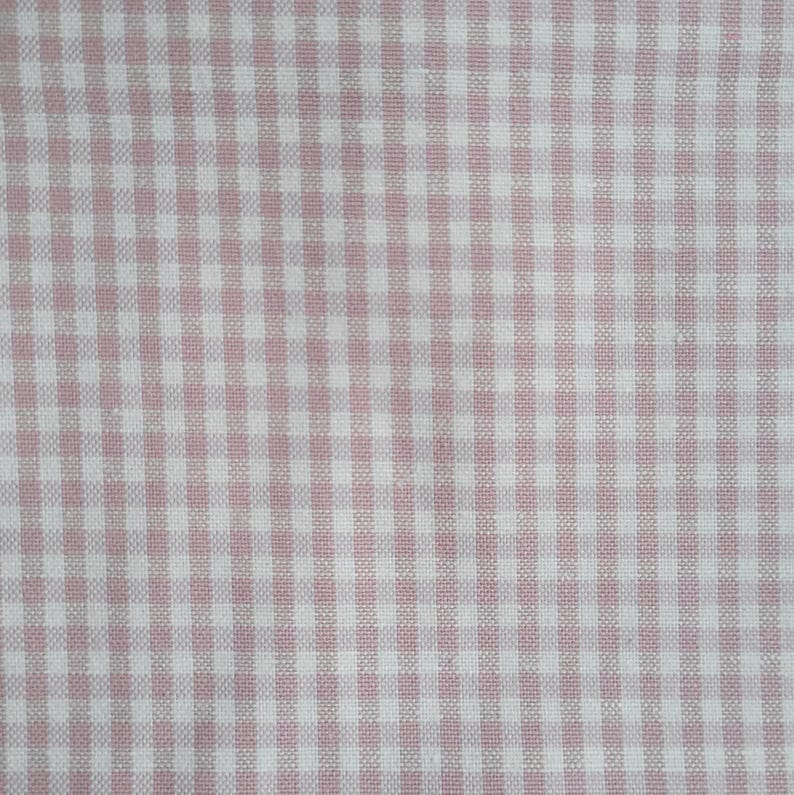 Pode incluir: Close-up de um tecido com padr&atilde;o xadrez vichy. O tecido apresenta uma grade repetida de pequenos quadrados em branco e rosa claro. O padr&atilde;o &eacute; uniformemente espa&ccedil;ado, criando um visual cl&aacute;ssico e texturizado.