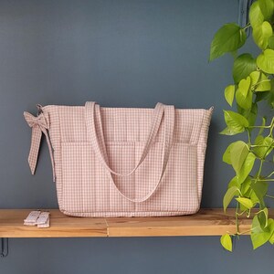 Pode incluir: Uma mala tote rosa e branca com padr&atilde;o xadrez, duas al&ccedil;as e bolsos exteriores. Uma fita a condizer est&aacute; presa na lateral. A mala est&aacute; numa prateleira de madeira.