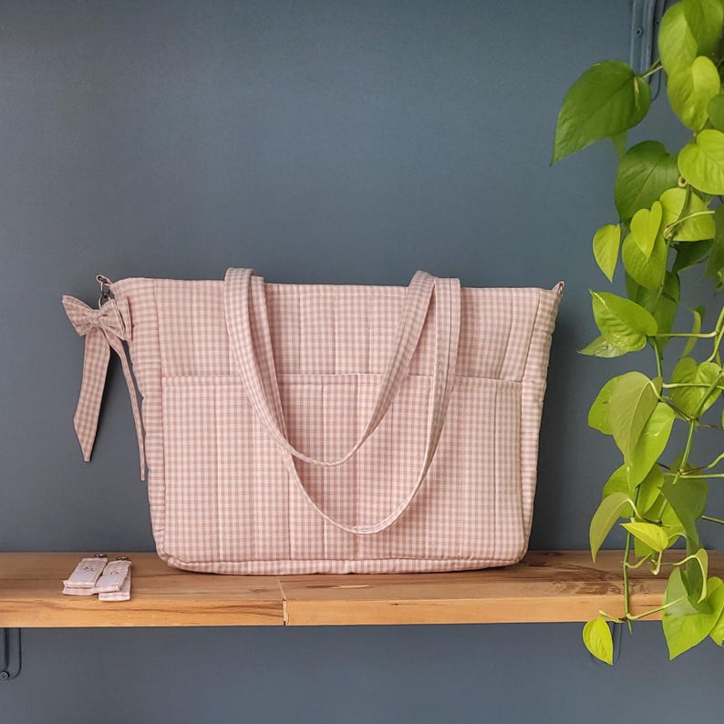 Pode incluir: Uma mala tote rosa e branca com padr&atilde;o xadrez, duas al&ccedil;as e bolsos exteriores. Uma fita a condizer est&aacute; presa na lateral. A mala est&aacute; numa prateleira de madeira.