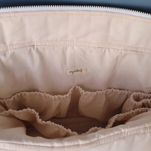 Pode incluir: Bolsa de fraldas de tecido bege com v&aacute;rios bolsos internos para organiza&ccedil;&atilde;o. A bolsa tem um z&iacute;per branco e uma pequena etiqueta que diz "mycalest". A bolsa est&aacute; aberta, revelando os compartimentos internos.