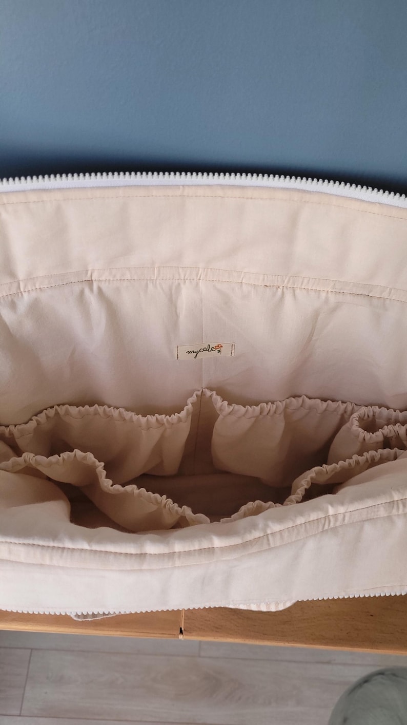 Pode incluir: Bolsa de fraldas de tecido bege com v&aacute;rios bolsos internos para organiza&ccedil;&atilde;o. A bolsa tem um z&iacute;per branco e uma pequena etiqueta que diz "mycalest". A bolsa est&aacute; aberta, revelando os compartimentos internos.