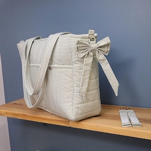 Pode incluir: Uma sacola tote xadrez verde claro e branco com al&ccedil;as combinando e um la&ccedil;o decorativo. A sacola tem dois bolsos externos e um fecho de z&iacute;per. As al&ccedil;as e o la&ccedil;o s&atilde;o feitos do mesmo tecido xadrez. A sacola est&aacute; em uma prateleira de madeira.