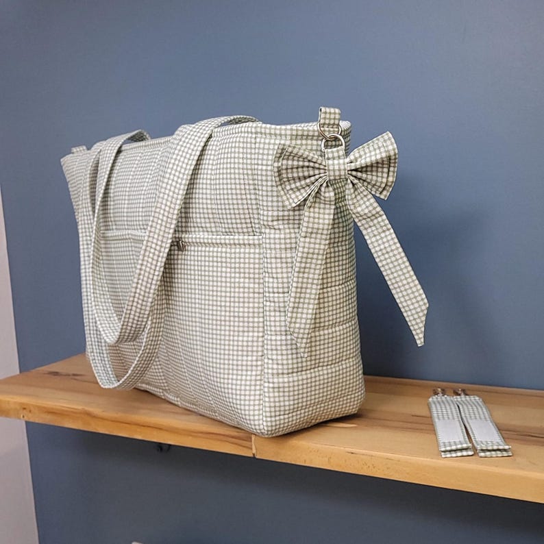 Pode incluir: Uma sacola tote xadrez verde claro e branco com al&ccedil;as combinando e um la&ccedil;o decorativo. A sacola tem dois bolsos externos e um fecho de z&iacute;per. As al&ccedil;as e o la&ccedil;o s&atilde;o feitos do mesmo tecido xadrez. A sacola est&aacute; em uma prateleira de madeira.