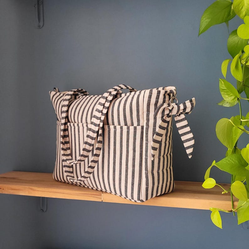 Pode incluir: Uma sacola listrada com listras verticais em preto e branco. A sacola tem dois bolsos externos e duas al&ccedil;as. A sacola est&aacute; em uma prateleira de madeira. A sacola tem uma fita lateral.