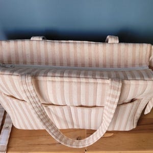 Pode incluir: Bolsa tote listrada bege e branca com v&aacute;rios bolsos e fecho de z&iacute;per. A bolsa tem duas al&ccedil;as e uma al&ccedil;a de ombro ajust&aacute;vel. O interior &eacute; vis&iacute;vel, mostrando um compartimento espa&ccedil;oso.