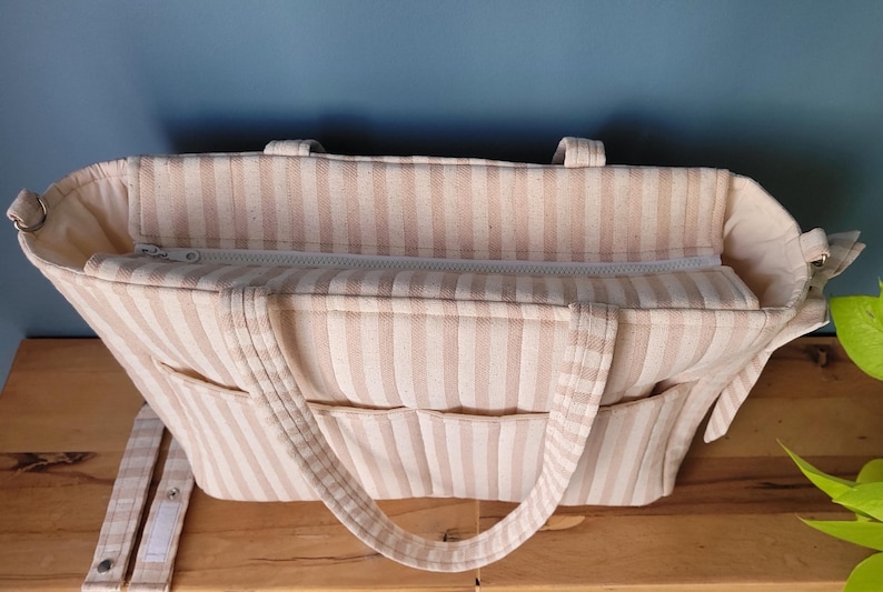 Pode incluir: Bolsa tote listrada bege e branca com v&aacute;rios bolsos e fecho de z&iacute;per. A bolsa tem duas al&ccedil;as e uma al&ccedil;a de ombro ajust&aacute;vel. O interior &eacute; vis&iacute;vel, mostrando um compartimento espa&ccedil;oso.