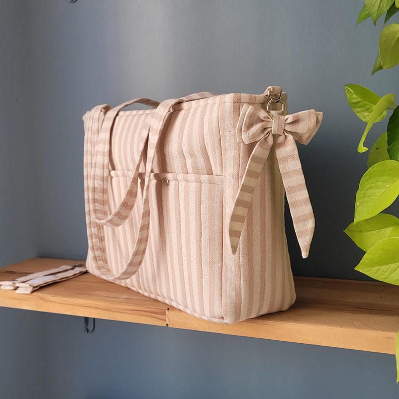 Pode incluir: Uma bolsa tote listrada bege e branca com duas al&ccedil;as e um la&ccedil;o decorativo. A bolsa tem dois bolsos externos e est&aacute; em uma prateleira de madeira. A bolsa &eacute; feita de um tecido macio e acolchoado.