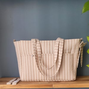 Pode incluir: Bolsa tote listrada bege e creme com duas al&ccedil;as e dois bolsos externos. A bolsa tem fecho de bot&atilde;o e um la&ccedil;o decorativo na lateral. Al&ccedil;as adicionais s&atilde;o vis&iacute;veis.