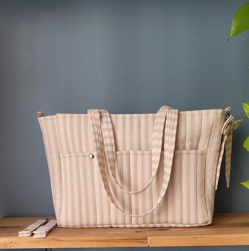 Pode incluir: Bolsa tote listrada bege e creme com duas al&ccedil;as e dois bolsos externos. A bolsa tem fecho de bot&atilde;o e um la&ccedil;o decorativo na lateral. Al&ccedil;as adicionais s&atilde;o vis&iacute;veis.