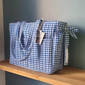 Pode incluir: Uma grande bolsa tote acolchoada com estampa xadrez vichy azul e branco. A bolsa tem dois bolsos externos, al&ccedil;as longas e um la&ccedil;o decorativo em um lado. Uma etiqueta est&aacute; presa &agrave; al&ccedil;a. A bolsa est&aacute; em uma prateleira de madeira.