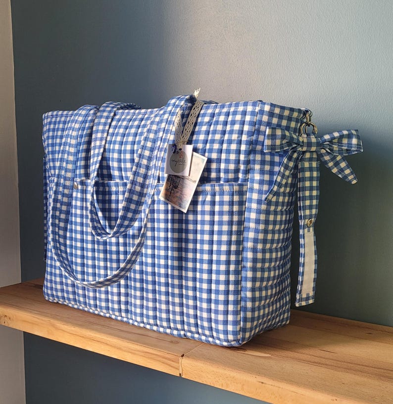 Pode incluir: Uma grande bolsa tote acolchoada com estampa xadrez vichy azul e branco. A bolsa tem dois bolsos externos, al&ccedil;as longas e um la&ccedil;o decorativo em um lado. Uma etiqueta est&aacute; presa &agrave; al&ccedil;a. A bolsa est&aacute; em uma prateleira de madeira.