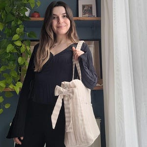 Pode incluir: Uma sacola tote listrada bege e branca com um grande la&ccedil;o decorativo. A sacola tem um fecho de cord&atilde;o e um bolso lateral. A sacola &eacute; segurada por uma pessoa. A sacola &eacute; feita de um tecido macio e texturizado.