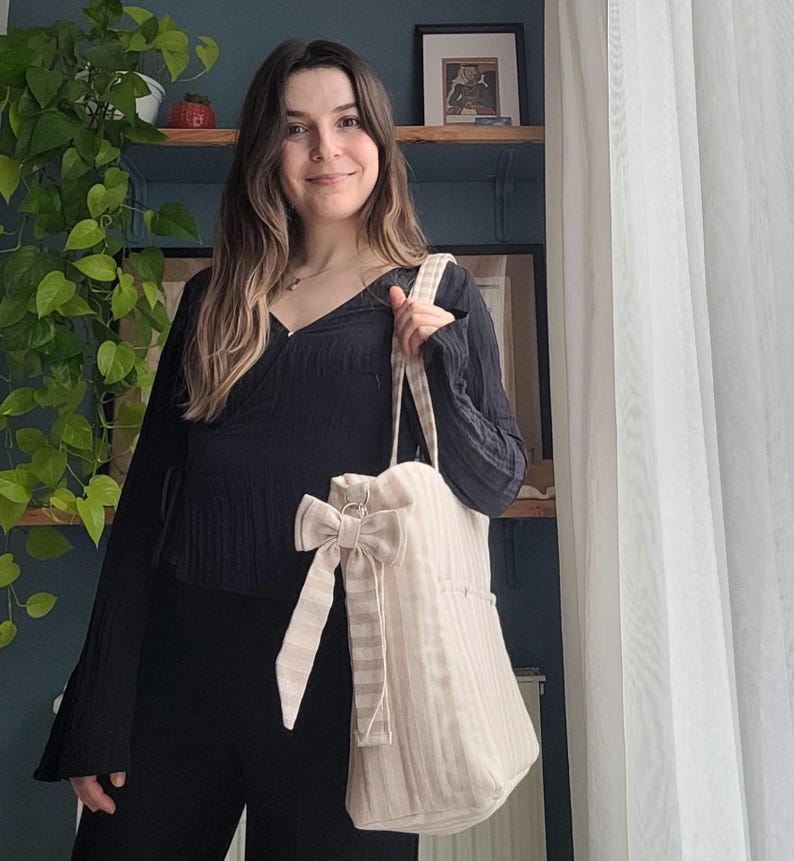 Pode incluir: Uma sacola tote listrada bege e branca com um grande la&ccedil;o decorativo. A sacola tem um fecho de cord&atilde;o e um bolso lateral. A sacola &eacute; segurada por uma pessoa. A sacola &eacute; feita de um tecido macio e texturizado.