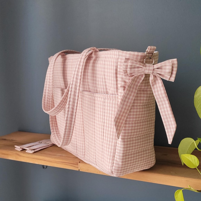Pode incluir: Bolsa tote rosa e branca com estampa xadrez, duas al&ccedil;as e uma al&ccedil;a de ombro. A bolsa tem dois bolsos externos e um la&ccedil;o decorativo. A bolsa est&aacute; em uma prateleira de madeira.